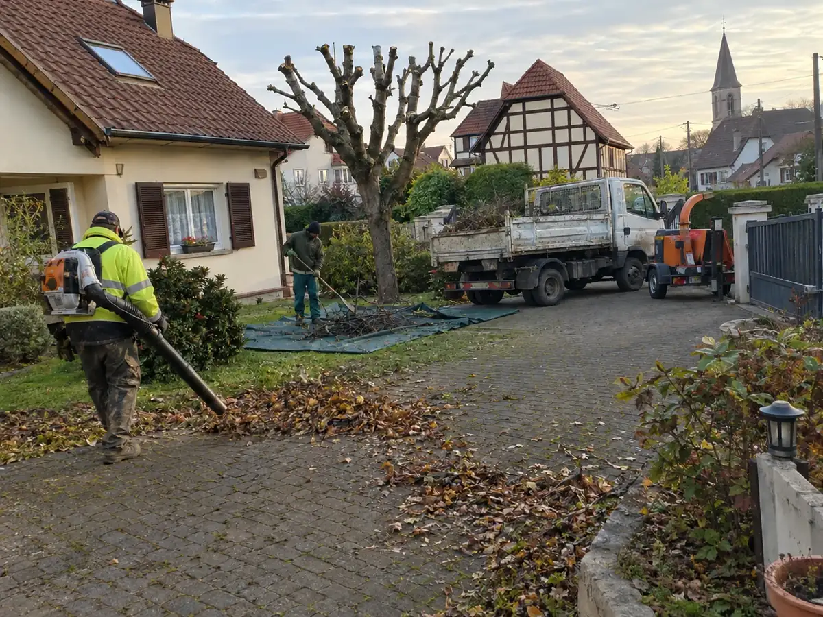 Fin de chantier : un ouvrier souffle les feuilles de l'allée, l'autre ratisse les branches sur une bâche, devant un arbre taillé
